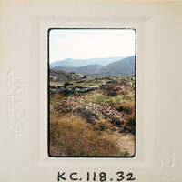 Photo of a mountainous landscape with a dirt road and scattered vegetation, presented as an old-fashioned film negative.