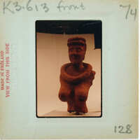A photograph of a clay figurine with a smiling face and arms crossed, displayed on a white surface.