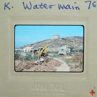 A photograph of a rural landscape with a dirt road, a building under construction, and a hillside.