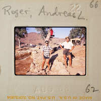 Two men standing on a rocky surface with mountains and trees in the background.