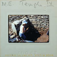 A person wearing a hat and a white shirt, standing next to a stone structure with a hole in the ground.