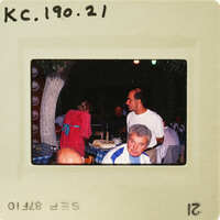 A vintage photograph of a group of people sitting at an outdoor dining table with a cake, showing signs of aging and wear. Catalog number KC.190.21