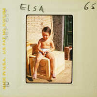 A young child sitting on a crate, looking down at their hands.