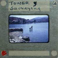 Man standing on rocks near water, with mountains and a village in the background.