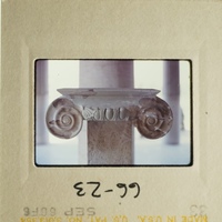 The photograph shows an ancient stone ionic column with decorative capitals and scrollwork, set against a plain background.