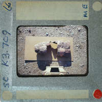 A photograph of an archaeological site with stone ruins and a measuring tool, taken from behind the camera.