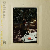 A vintage photograph of a woman sitting at an outdoor table, with a cat nearby and a dog in her lap.