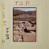 Ancient ruins with stone foundations and walls, set against a backdrop of rugged terrain and hills.