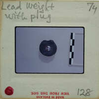 A small metal object with a hole in the center, photographed on a white surface with measurement lines and labels.