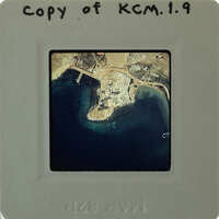 Aerial photograph of a coastal area with water and land features, enclosed by a white border on a film strip.