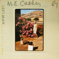 A vintage photo of a woman standing among pink flowers with mountains in the background.