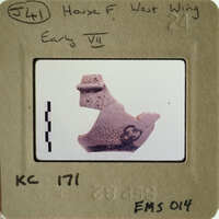 Ancient artifact on display, possibly a clay figurine with markings, photographed against a white background.