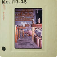 A vintage photograph of a rustic room with shelves, chairs, and a wheelbarrow.