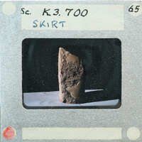 The image is a photograph of an old, weathered object with a label "SKIRT" on it, placed on a surface and surrounded by darkness.