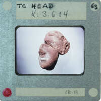 A close-up photograph of a small red figurine with a face, displayed on a white background.