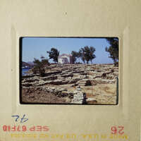 The photo shows an ancient Greek temple on a rocky hillside, with a clear blue sky and a few trees in the background.