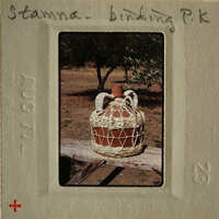 An old-fashioned clay jug with woven handles and body, sitting on a wooden surface outdoors.