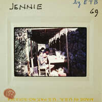 Old photograph of two people sitting at a table with a stone structure behind them, enclosed by a frame and labeled "JENNIE".