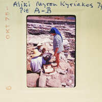 A vintage photo of a woman standing behind two men working on stone ruins, with handwritten notes and a stamp indicating it's from Ali Myron Kyriakos.
