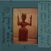 A clay figurine of a person with raised hands, displayed behind glass.