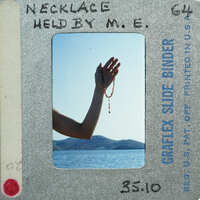 A hand holding a rosary beads against a scenic background with mountains and water.