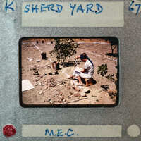 A man sitting on a stool with his legs crossed, reading a book next to a small tree and surrounded by dirt.