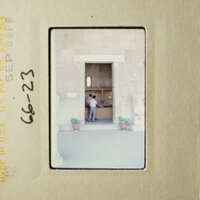 This is an old photograph of a person standing outside a building, possibly at a doorway or entrance, with another person inside.