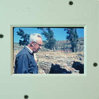 An old man wearing glasses and a jacket standing outside near stone ruins.
