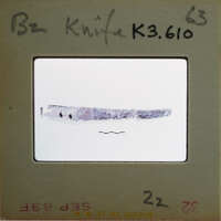 A small, curved metal object with a pointed end and a handle, possibly a knife or tool, on display in a case labeled "K360.610".