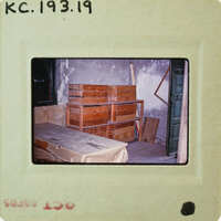Old photograph of a room with wooden crates and a bed frame, taken from an antique camera negative.