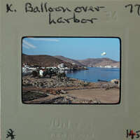 Photo of a harbor with mountains and sea, with balloon in the air.