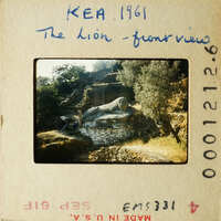 Photo of lion statue on stone base with trees and mountains in background.