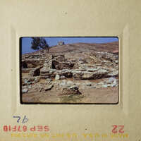 The image shows an ancient stone structure, possibly ruins, with a clear sky and vegetation in the background.