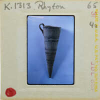 The image shows a cylindrical rhyton vase (K.1313) with a handle, displayed against a blue background.