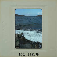 A vintage photo of a rocky shoreline with waves and an island in the background.