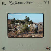 A photograph of a group of people standing near trees and rocks, with mountains in the background.