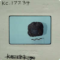 A small, dark rock specimen on a white surface with measurement lines.