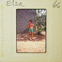 A young boy standing on dirt ground with a plant behind him, wearing blue shorts and no shirt.