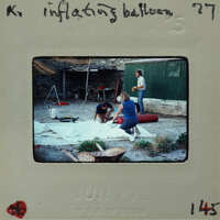 A vintage photograph of three people working on an inflatable object, with a handwritten note and date on the bottom.