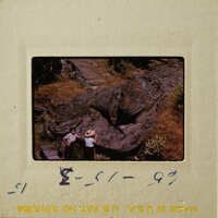 Photograph of two people standing near a large rock formation with vegetation around it.