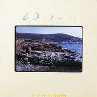 A photograph showing a rocky shoreline with water and distant hills.