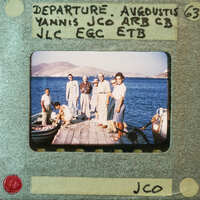 Old photograph of a group of people on a dock by the sea with mountains in the background.