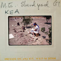 Person sitting on a stool outdoors, working with clay and pottery tools.