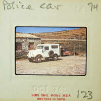 A vintage photo of a police car parked next to a stone building with mountains in the background.