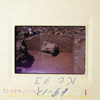 A photograph of an archaeological site with stone ruins, taken from a high angle and showing a person standing on a mound of earth.