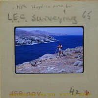 A man standing on a cliff overlooking the ocean, with mountains in the background.