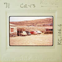 Old cars parked outside a stone building with mountains in the background.