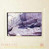 The photo shows an archaeological site with stone ruins and a small stream, taken from a high angle perspective.