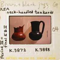 Two neck-handled tankards, one brown and the other black, displayed on a table with labels.
