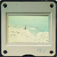 A vintage photograph of a person standing on rocks by the ocean, encased in an old-fashioned frame with visible screws and numbers indicating its age.